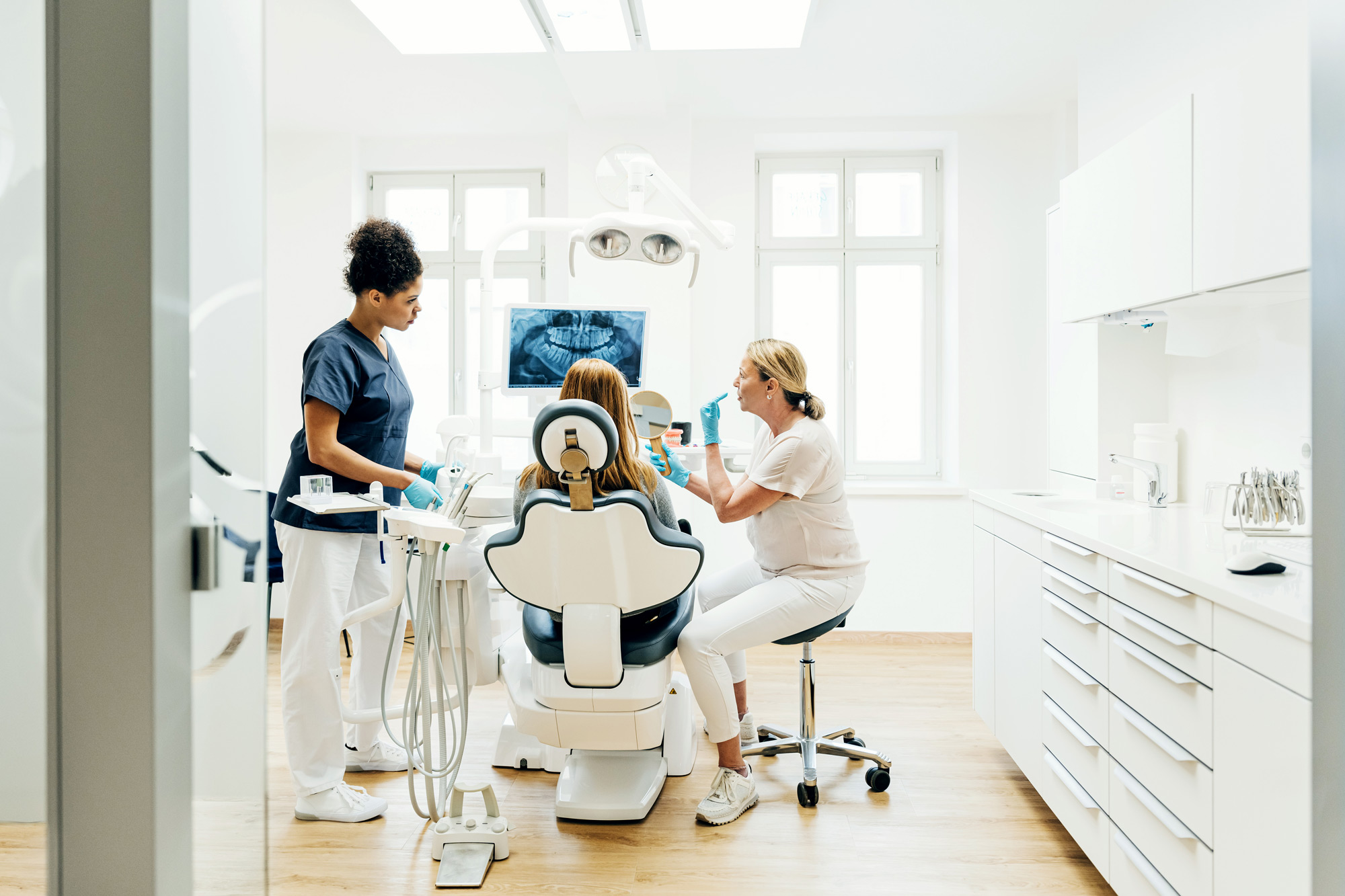 En tandläkare i marinblå sjukhuskläder visar en röntgenbild av tänder på en datorskärm till en ung patient som sitter i tandläkarstolen.